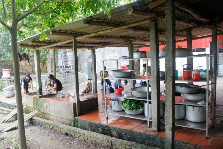 Khu vực nấu ăn ngoài trời có mái che với những người đang chuẩn bị thực phẩm bằng chậu và nồi kim loại lớn, cây xanh xung quanh, trời u ám